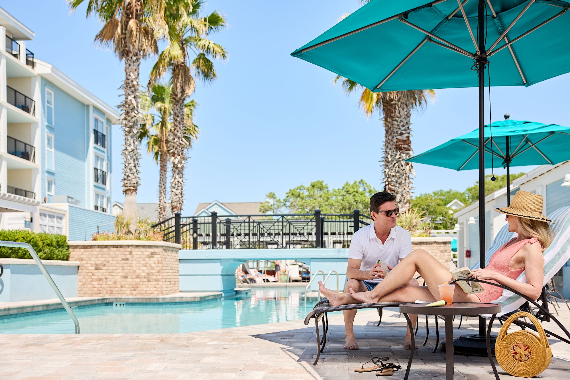 Two people are lounging by a pool under a blue umbrella, enjoying a sunny day with drinks, surrounded by palm trees and buildings.