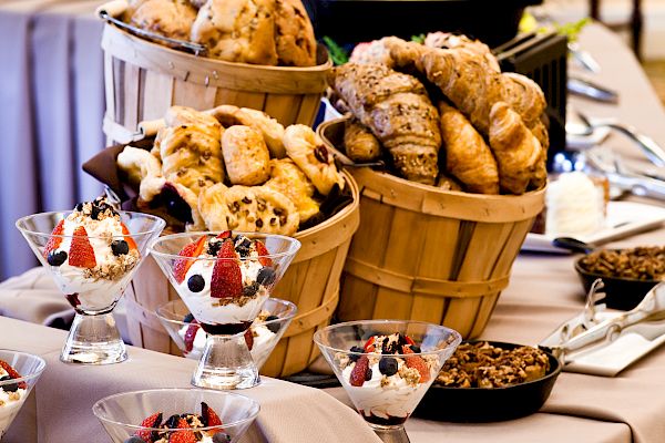 A buffet table with baskets of bread, bowls of fruit, and various dishes elegantly arranged on a table with a beige cloth.