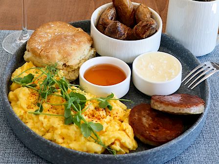 A breakfast platter with scrambled eggs, biscuit, sausage, gravy, potato wedges, syrup, coffee, and a mimosa on the side.