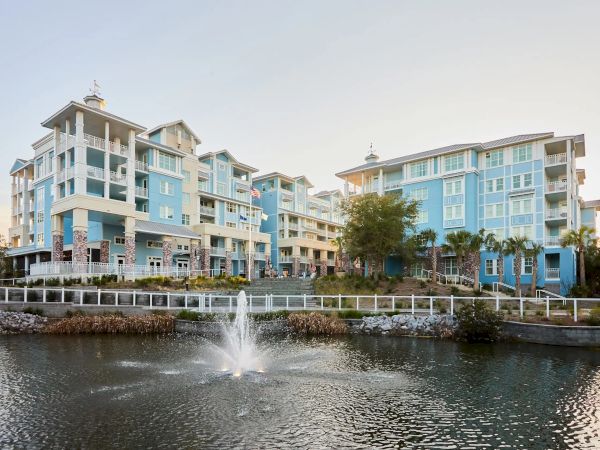 A group of blue-and-white buildings with a fountain in a pond in front, surrounded by a fence and trees, under a clear sky.