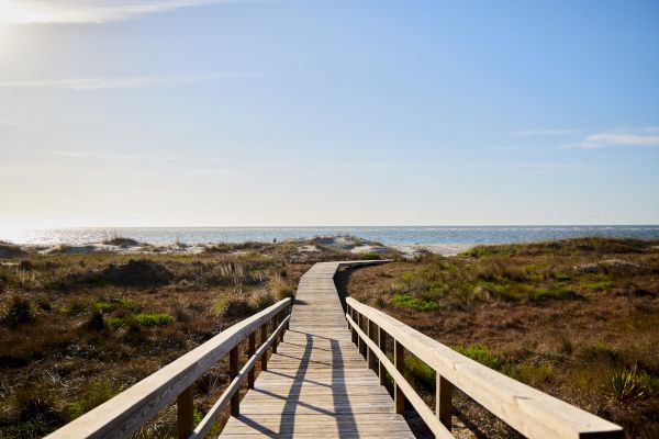 A wooden boardwalk leading over dunes to a calm, sunlit beach with clear skies and grassy vegetation.