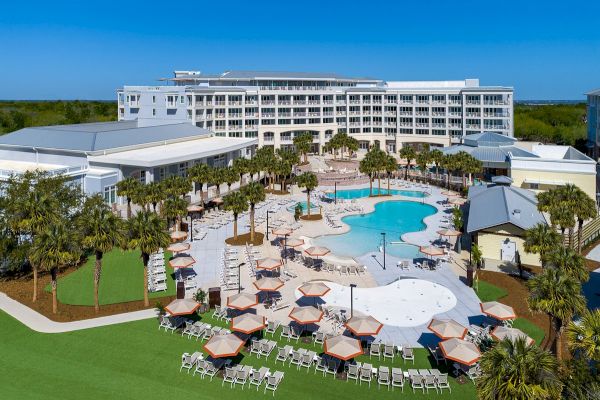 The image shows a resort with a large pool, surrounding loungers, umbrellas, and palm trees, adjacent to a multi-story building.