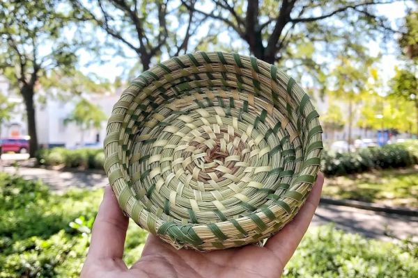 A person is holding a small, woven basket outside with trees and greenery in the background.