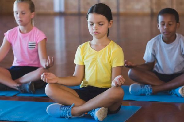 Children sitting cross-legged on yoga mats, eyes closed, practicing meditation in a gymnasium setting.