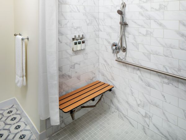 A bathroom shower with a foldable wooden bench, a wall-mounted showerhead, grab bar, and toiletries on a tiled wall.