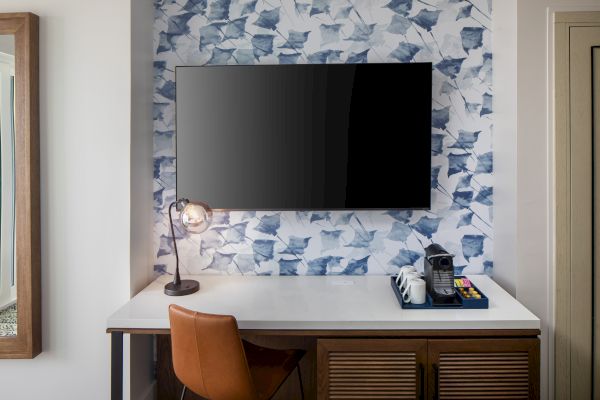 A modern desk setup with a TV, lamp, coffee maker, mugs, and snacks on a patterned wall backdrop.