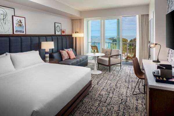 A modern hotel room with a bed, sofa, chairs, desk, and a balcony with an ocean view. Artwork decorates the walls.