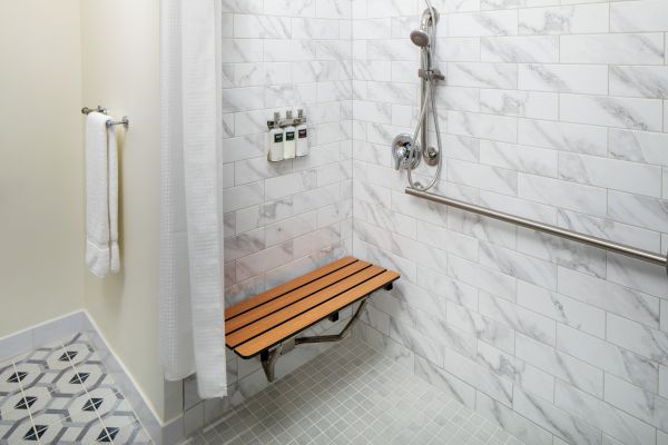 This image shows a shower with a fold-down bench, a handheld showerhead, grab bar, and wall-mounted soap dispensers, and a towel hanging nearby.