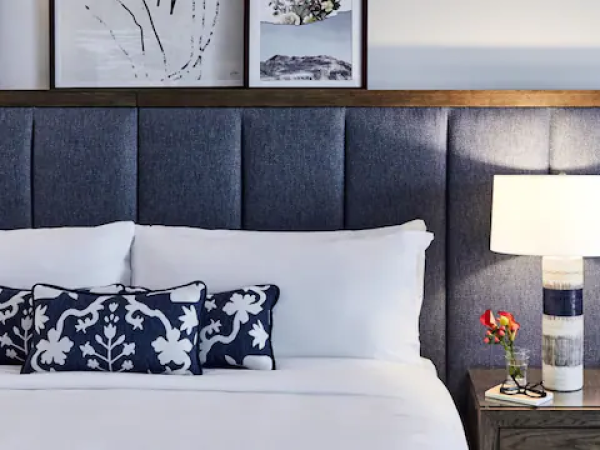 A bed with blue floral pillows sits against a padded headboard. A lit lamp and flowers are on a wooden nightstand, with art above.