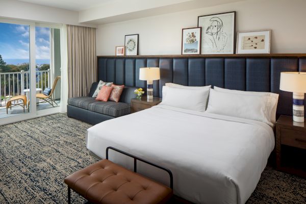 Modern hotel room with a king bed, lamps, artwork, and a seating area by a window leading to a balcony with chairs and a table.