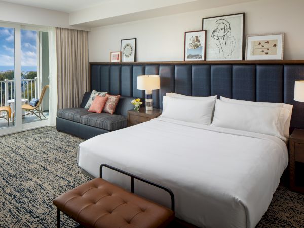 Modern hotel room with a king bed, lamps, artwork, and a seating area by a window leading to a balcony with chairs and a table.