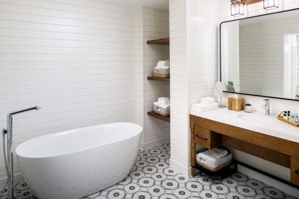 A modern bathroom with a freestanding bathtub, wooden vanity, large mirror, geometric floor tiles, and shelves with towels and toiletries.