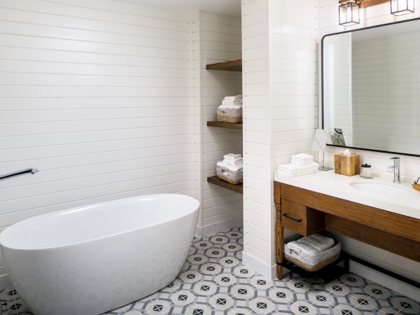 A modern bathroom with a freestanding bathtub, wooden vanity, large mirror, geometric floor tiles, and shelves with towels and toiletries.
