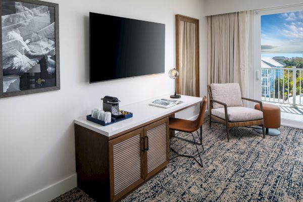 A modern hotel room with a TV, desk, coffee maker, lamp, chair, and a view of a balcony with blue skies.