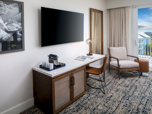 A modern hotel room with a TV, desk, coffee maker, lamp, chair, and a view of a balcony with blue skies.