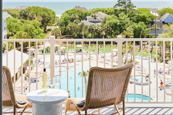 A balcony with two chairs and a table with wine, overlooking a pool and lush greenery leading to the ocean.