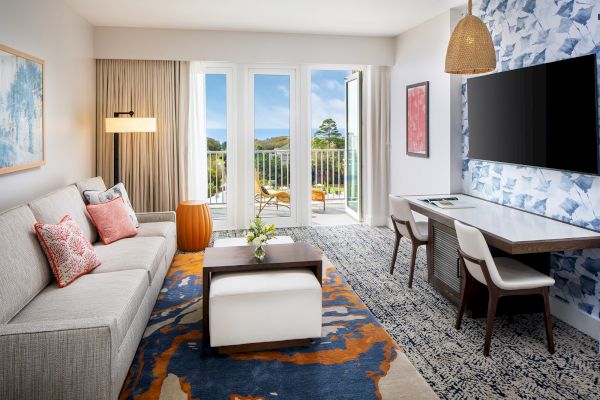 Modern living room with a sofa, colorful pillows, coffee table, TV, desk, chairs, and balcony view. Decorated with art and rug patterns.