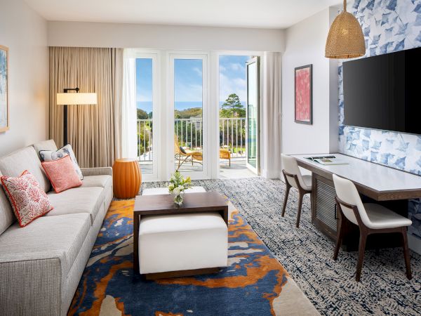 Modern living room with a sofa, colorful pillows, coffee table, TV, desk, chairs, and balcony view. Decorated with art and rug patterns.