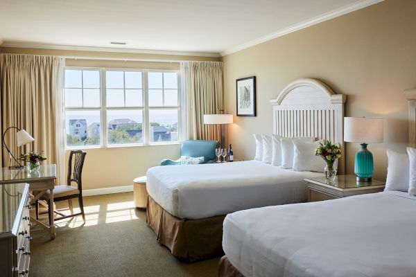 A hotel room with two beds, large window, chair, lamps, wine, and coastal view. Decor includes flowers and framed art on the wall.