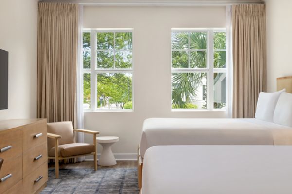 The image shows a hotel room with two beds, a TV, a wooden dresser, a chair, and large windows with a view of greenery outside.