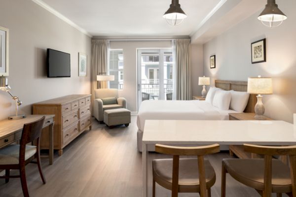 A modern hotel room with double beds, a TV, desk, chairs, lamps, and a balcony view. Neutral colors create a calm atmosphere.