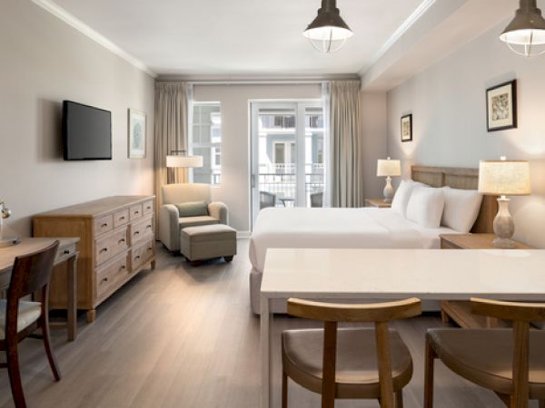 A modern hotel room with double beds, a TV, desk, chairs, lamps, and a balcony view. Neutral colors create a calm atmosphere.