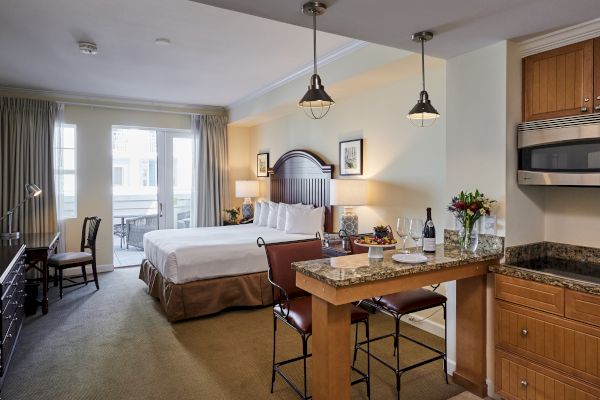 A cozy hotel room with a bed, desk, kitchen area, and a small dining counter; doors lead to a balcony, and flowers add a touch.