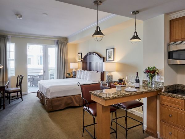 A cozy hotel room with a bed, desk, kitchen area, and a small dining counter; doors lead to a balcony, and flowers add a touch.