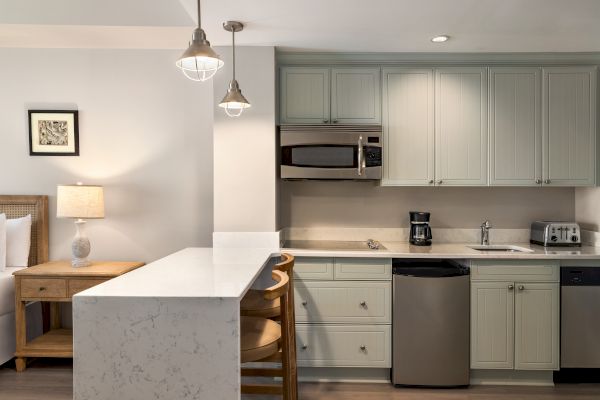 A modern kitchen with light cabinetry, countertop seating, stainless steel appliances, hanging lights, and a cozy side table with a lamp.