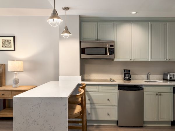 A modern kitchen with light cabinetry, countertop seating, stainless steel appliances, hanging lights, and a cozy side table with a lamp.