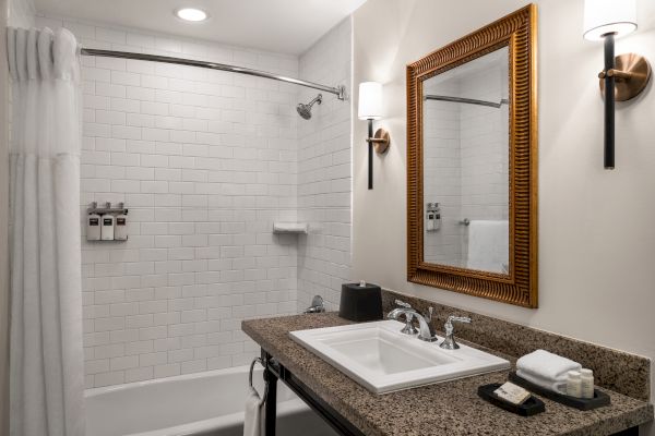 A modern bathroom with a shower, white tiles, a granite countertop, sink, mirror, and wall-mounted soap dispensers and lighting.