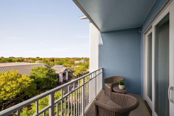 A balcony with wicker chairs and a small table overlooks a green, tree-filled area with buildings and a clear sky.