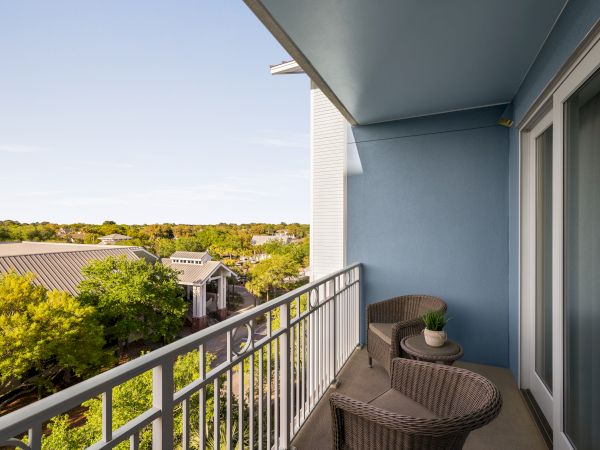 A balcony with wicker chairs and a small table overlooks a green, tree-filled area with buildings and a clear sky.