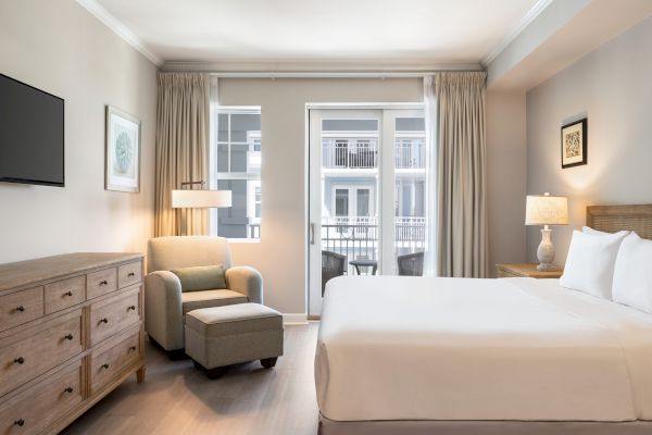 The image shows a modern hotel room with a bed, armchair, ottoman, dresser, lamps, and a balcony view.