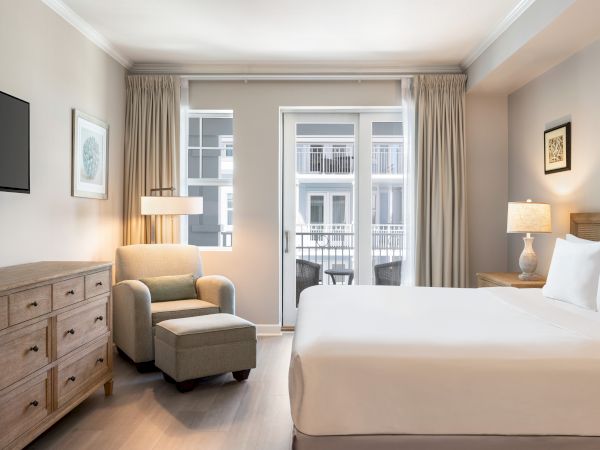 The image shows a modern hotel room with a bed, armchair, ottoman, dresser, lamps, and a balcony view.