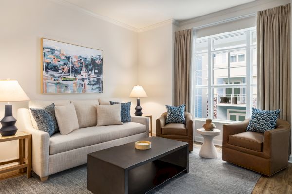 A modern living room with a couch, two chairs, lamps, artwork, and a coffee table near a large window with curtains.