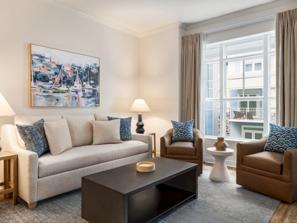 A modern living room with a couch, two chairs, lamps, artwork, and a coffee table near a large window with curtains.
