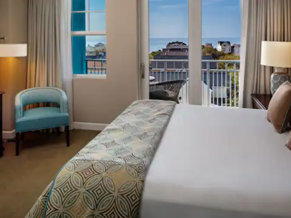 A cozy hotel room features a bed, TV, and armchair. Large windows offer a view of buildings and the sea beyond the balcony.
