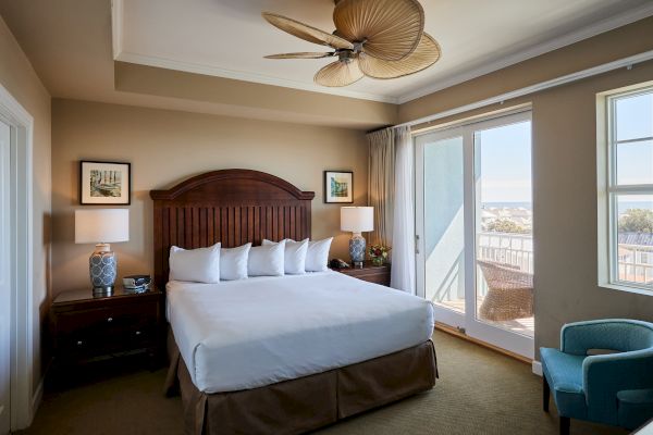A neatly arranged bedroom with a wooden bed, white bedding, lamps, artwork, a blue chair, and a balcony view through glass doors.