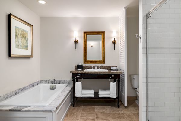 A modern bathroom with a bathtub, framed art, sink, mirror, lights, towels, and a shower area is shown.