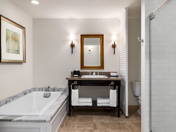 A modern bathroom with a bathtub, framed art, sink, mirror, lights, towels, and a shower area is shown.