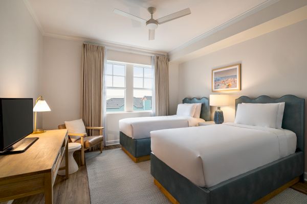 The image shows a hotel room with two beds, a desk, chair, lamp, and TV, with a window and decorative artwork on the wall.