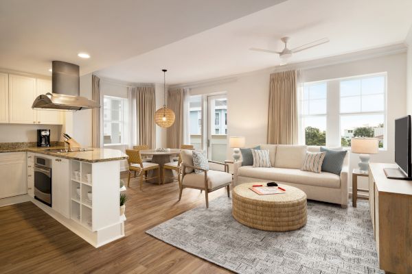 A modern open-plan living area with a kitchen, dining set, sofa, TV, and decor, featuring light colors and wood flooring.