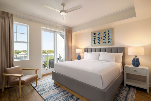 A modern bedroom with a grey bed, white bedding, blue lamps, art on the wall, and a balcony view.