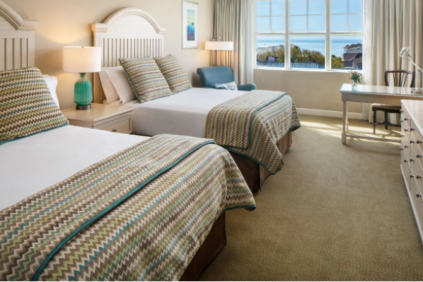 The image shows a hotel room with two beds, rattan headboards, a TV, a desk, a chair, and a window with a view of trees and the ocean.