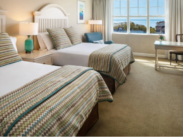 The image shows a hotel room with two beds, rattan headboards, a TV, a desk, a chair, and a window with a view of trees and the ocean.