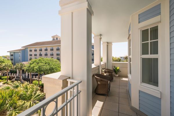 A sunny balcony with white columns, wicker chairs, and a potted plant overlooks a landscaped area with trees and a multi-storied building.