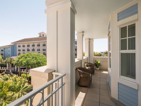 A sunny balcony with white columns, wicker chairs, and a potted plant overlooks a landscaped area with trees and a multi-storied building.