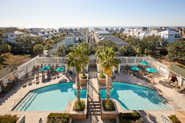 A pool area with palm trees and sun loungers is surrounded by houses, with a view extending to a coastal horizon.