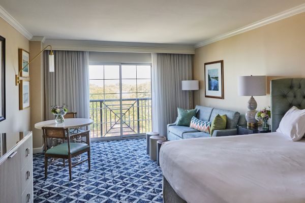 A well-lit hotel room with a bed, sitting area, table, and a view through a large window. Decor includes lamps and framed art.